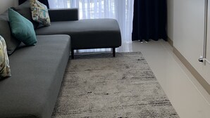 Un salon avec un canapé gris, des coussins turquoise, un pouf noir, un tapis gris, un téléviseur à écran plat fixé au mur et un balcon avec des rideaux blancs.