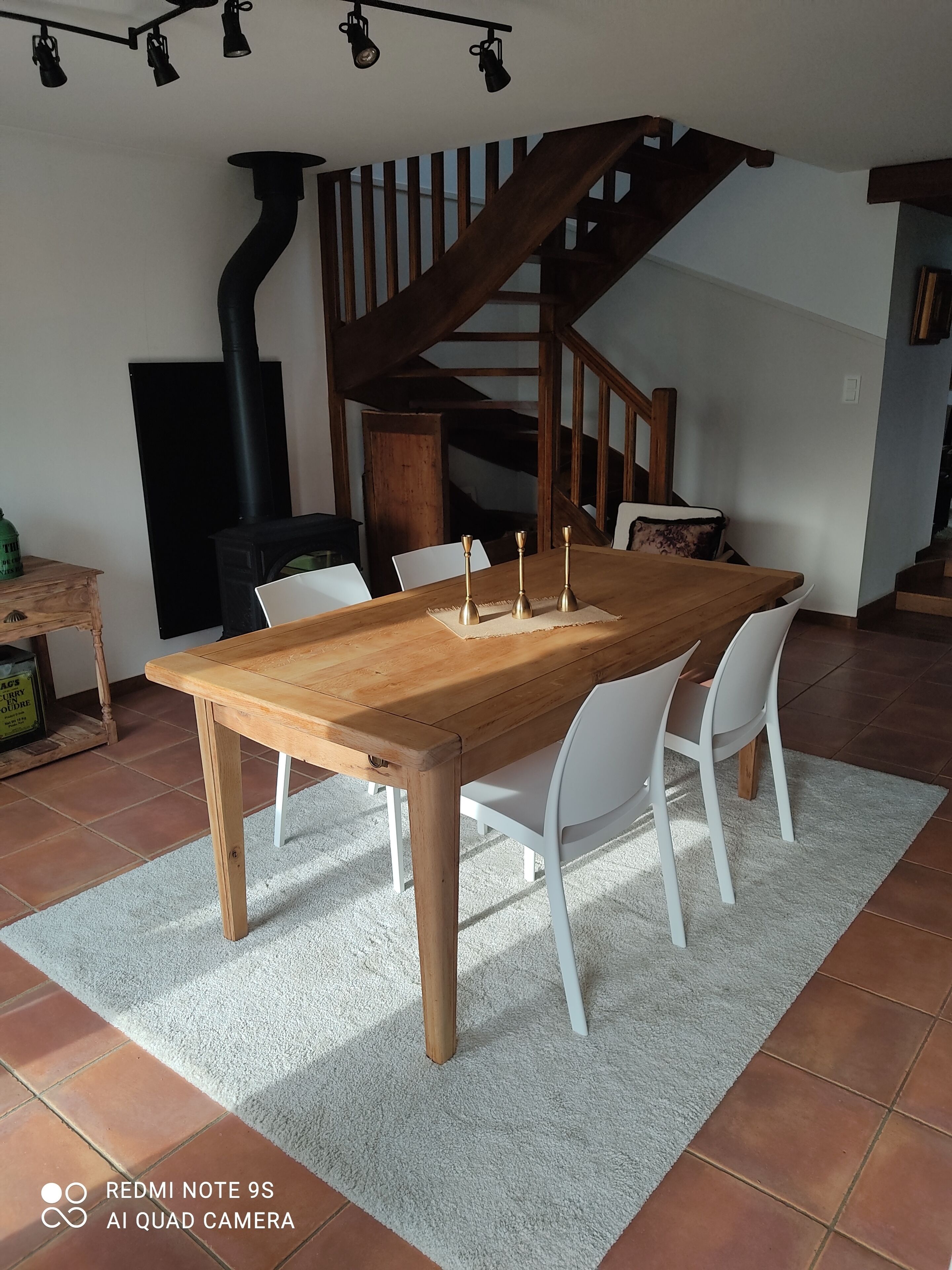A dining area with a wooden table, white chairs, a white rug, and a staircase in the background.
