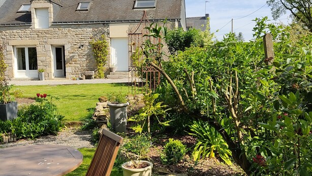 A stone house with a sloped roof, a small front porch, and a well-maintained garden with various plants and a wooden chair. - Country house on the edge of a forest with landscaped garden. (Billio)