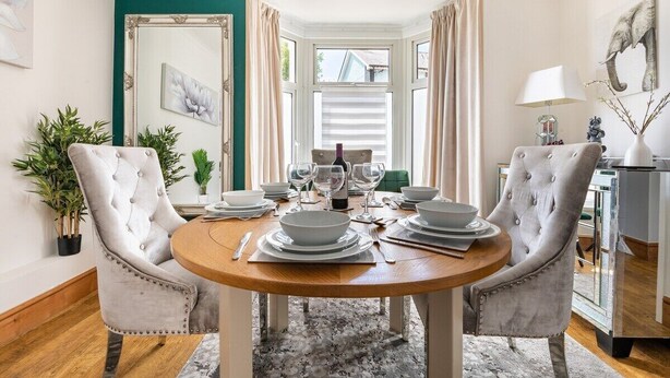 A dining area with a round wooden table set for four, surrounded by tufted chairs. There is a large mirror on the wall, a potted plant, and a view of the outdoors through a window with curtains. - Ty Hapus is a modern, spacious townhouse featuring a King bed, fully equipped kitchen, and a private fenced backyard with a fire pit. Located in a highly walkable neighborhood near historic sites, vibrant dining, and beautiful beaches, room for six (Bridgend County Borough)