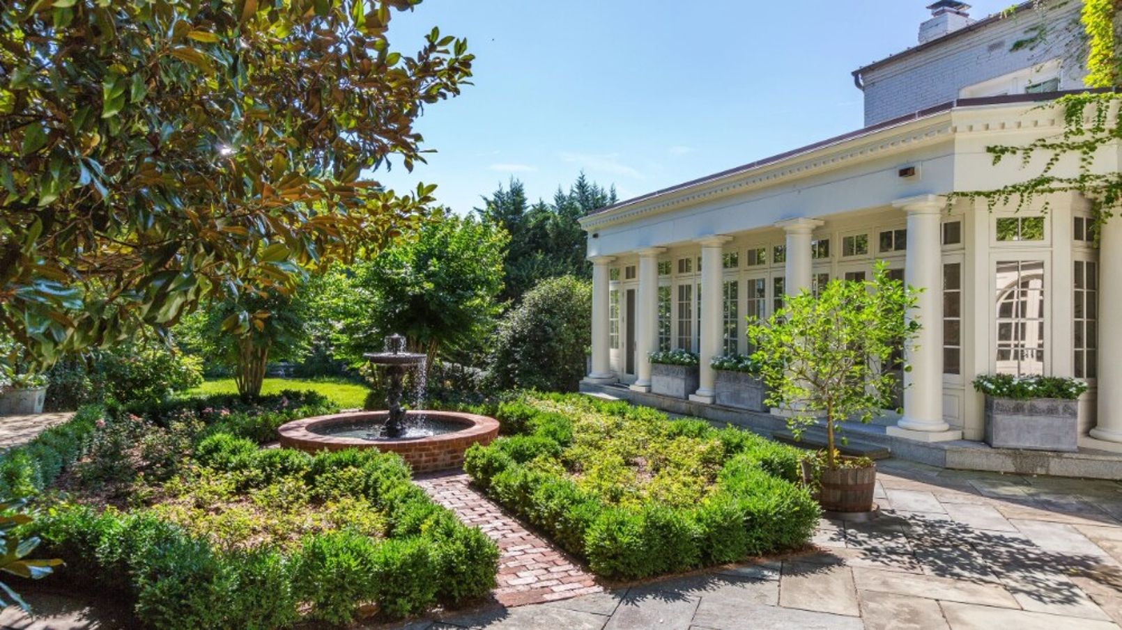 A formal garden with a fountain, neatly trimmed hedges, and a classical building with columns.