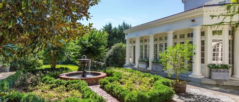 Ein formeller Garten mit Brunnen, ordentlich geschnittenen Hecken und einem klassizistischen Gebäude mit Säulen.
