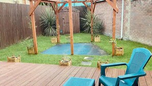 Una terraza de madera con un pérgola, dos sillas de plástico azules y una mesa pequeña.