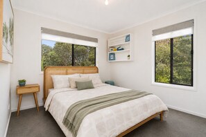 A bedroom with a bed, a bedside table, a wall-mounted shelf, and two windows with blinds.