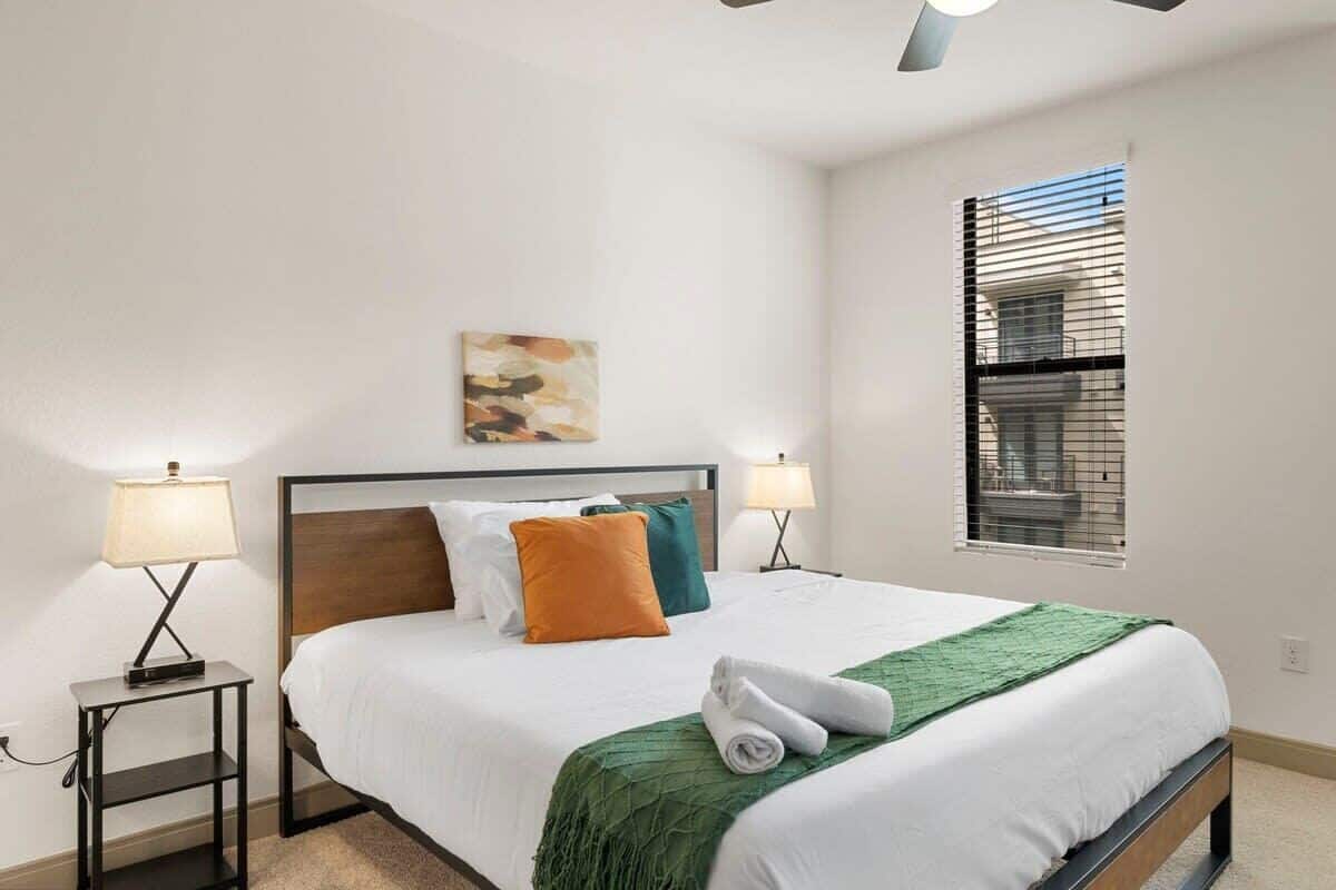 A hotel room with a bed, two bedside lamps, a window with blinds, and a painting on the wall.