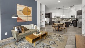 Ein modernes Wohnzimmer mit einer Couch, Couchtisch und einem gemusterten Teppich. Eine Küche mit weißen Schränken und ein Essbereich mit Tisch und Stühlen sind ebenfalls zu sehen.