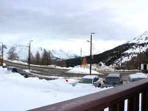 Parque de estacionamento coberto de neve, com carros e montanhas ao fundo.