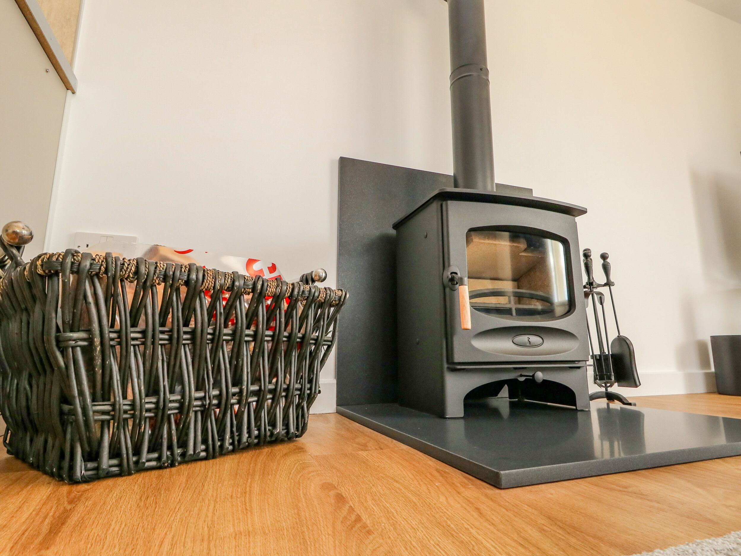 Een moderne kamer met een zwart houtkacheltje, een rieten mand met spullen en een houten vloer.