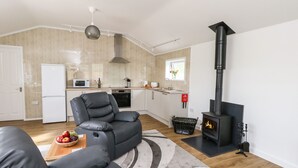 A modern living room with a grey sofa, a small wooden table with fruit, a black fireplace, and a kitchen in the background. - Cliff Barn (Bridport)
