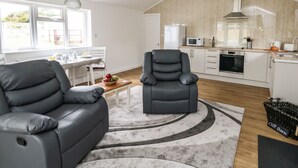 A modern living room with a grey sofa and armchair, a dining area with a table and chairs, and a kitchen with white cabinets and appliances. - Cliff Barn (Bridport)