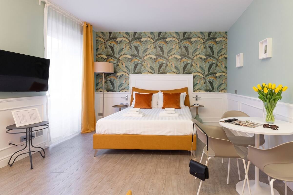 A hotel room with a bed, a desk, a chair, a TV, and a wall with a floral pattern.