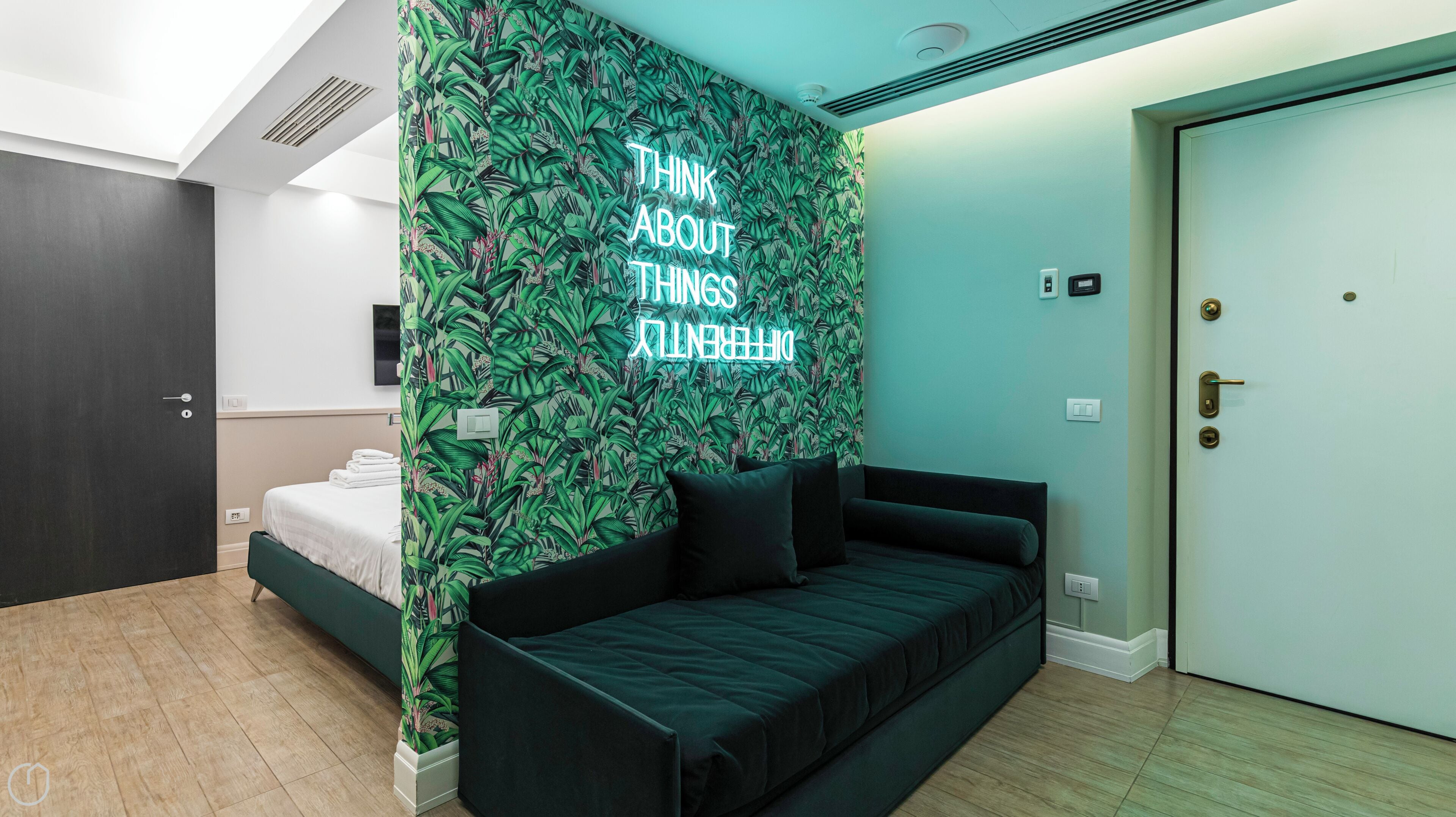 A modern hotel room with a bed, a dark green sofa, a neon sign on the wall, and a light blue door.