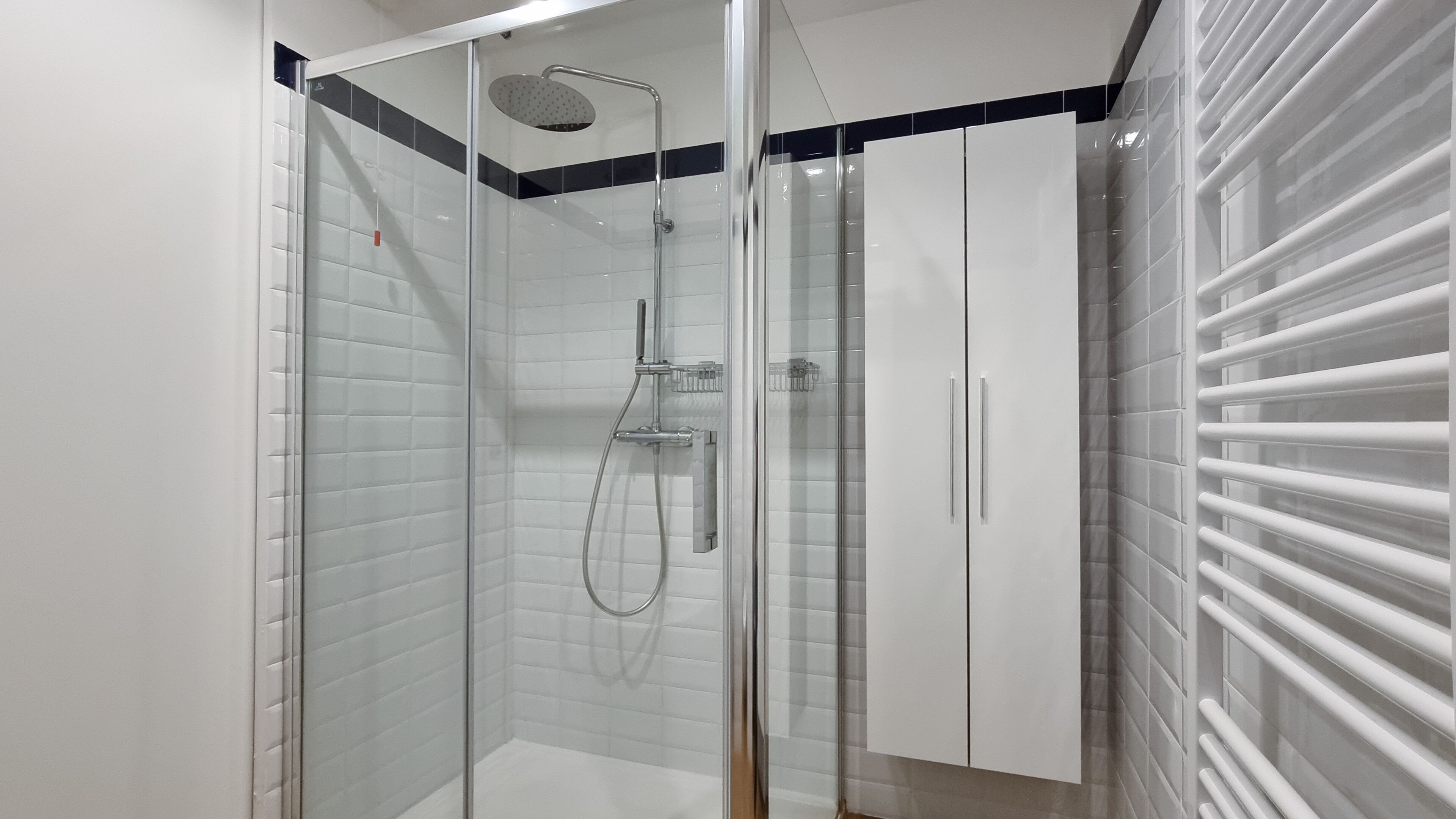 Um banheiro moderno com chuveiro envidraçado, paredes azulejadas em branco e um armário branco com faixa preta.