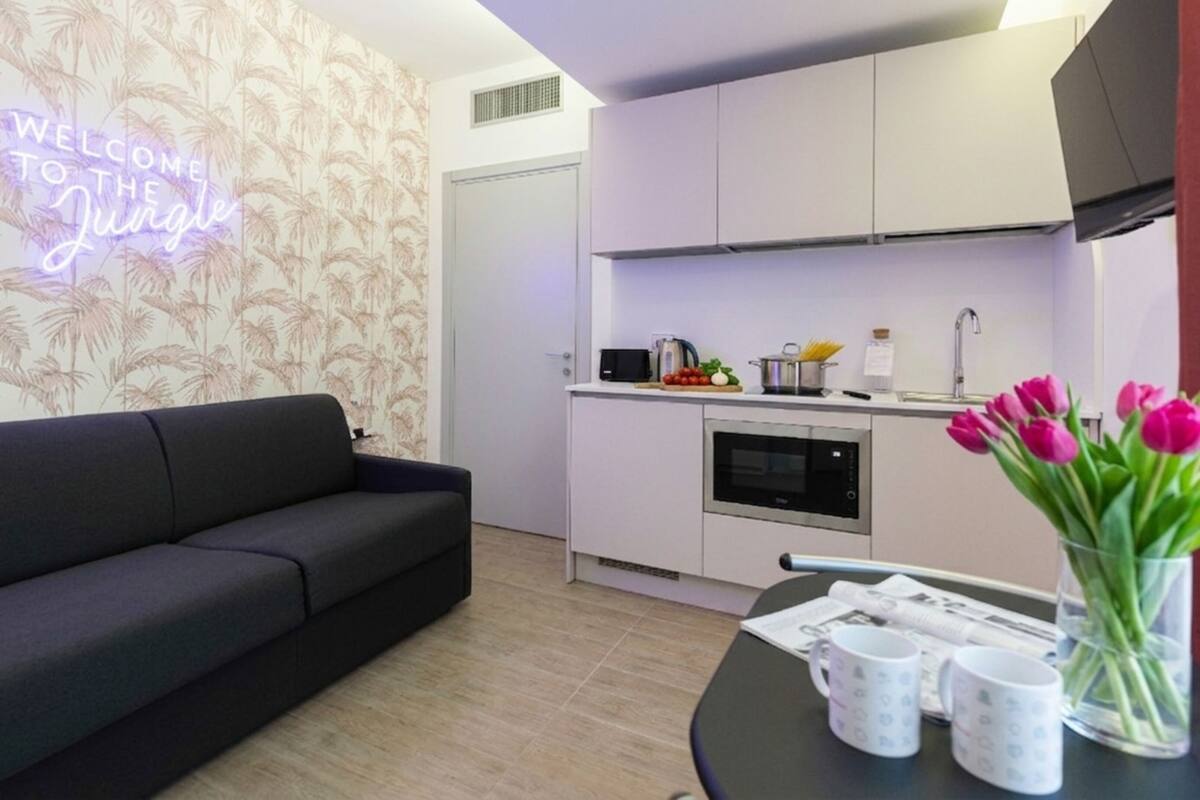 A modern living space with a sofa, a kitchen with appliances, and a neon sign on the wall.