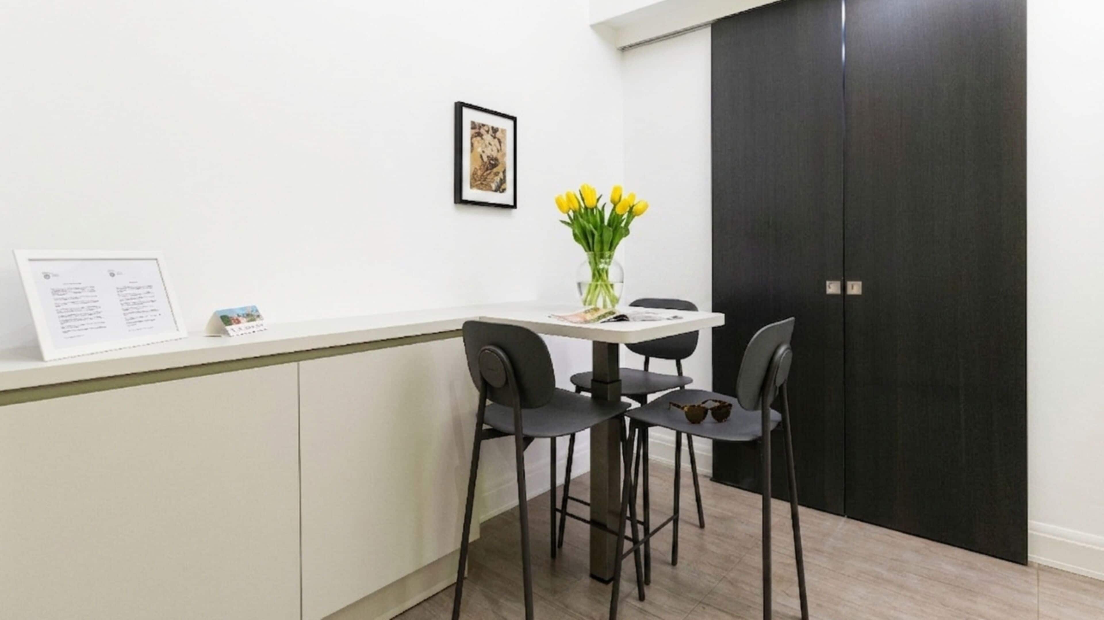 A small, modern kitchen with a white countertop, black chairs, and a vase with yellow flowers.