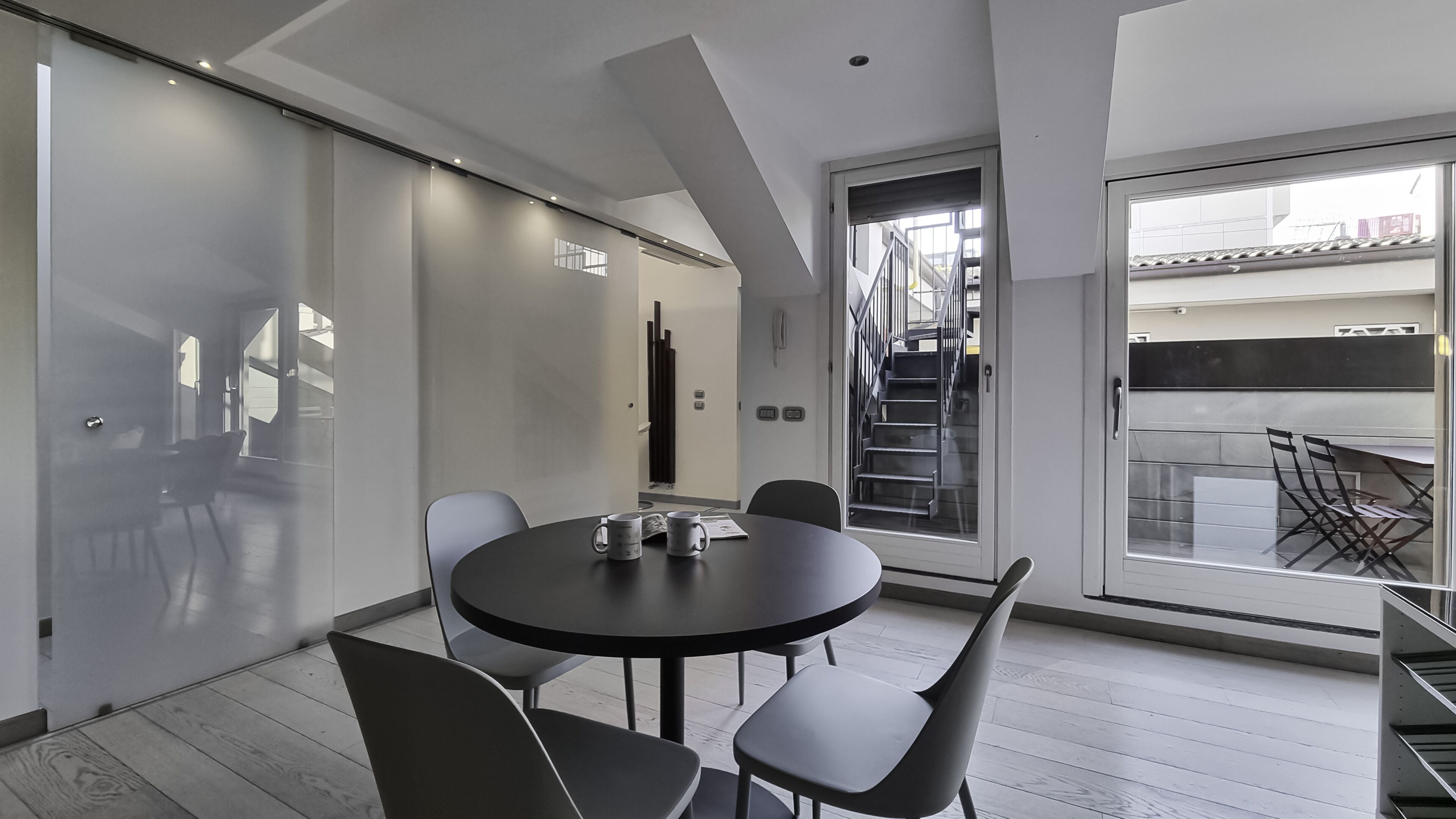 A modern dining area with a round table and chairs, a glass partition, and a view of a staircase and balcony.