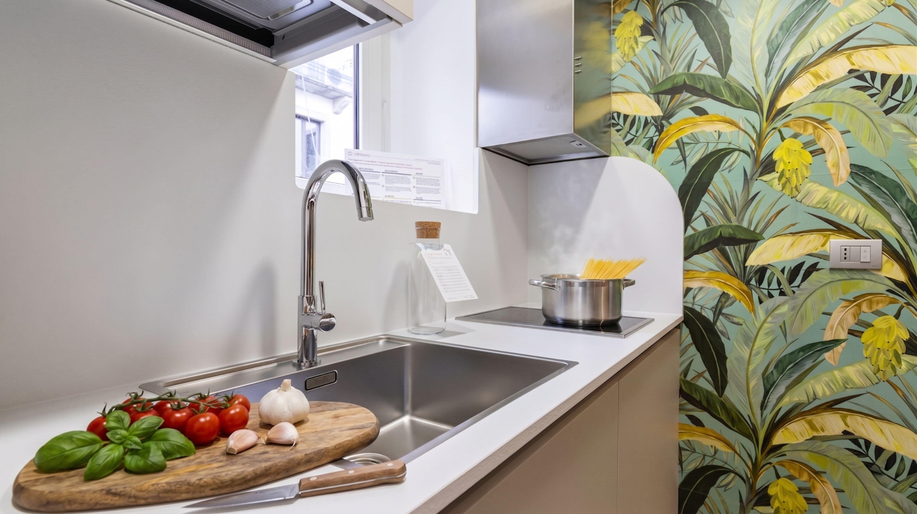 A kitchen with a stainless steel sink, a wooden cutting board with tomatoes and basil, and a pot on the countertop.