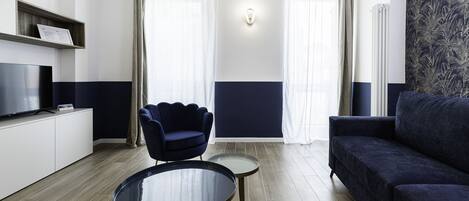 A modern living room with a flat-screen TV, a dark blue sofa, a matching armchair, and a small round table.