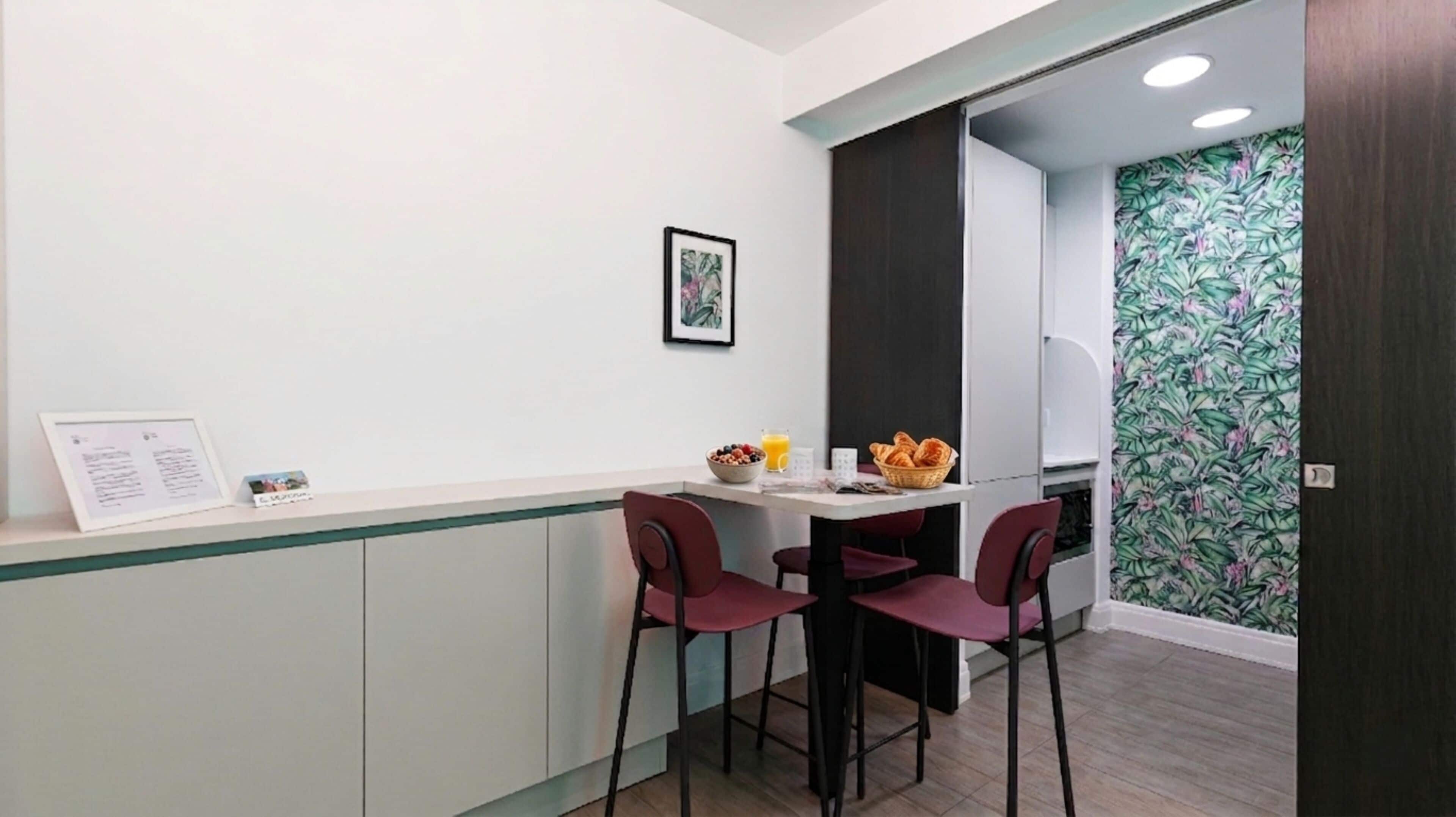 A small reception area with a counter, two red chairs, a framed picture on the wall, and a door with a floral wallpaper.