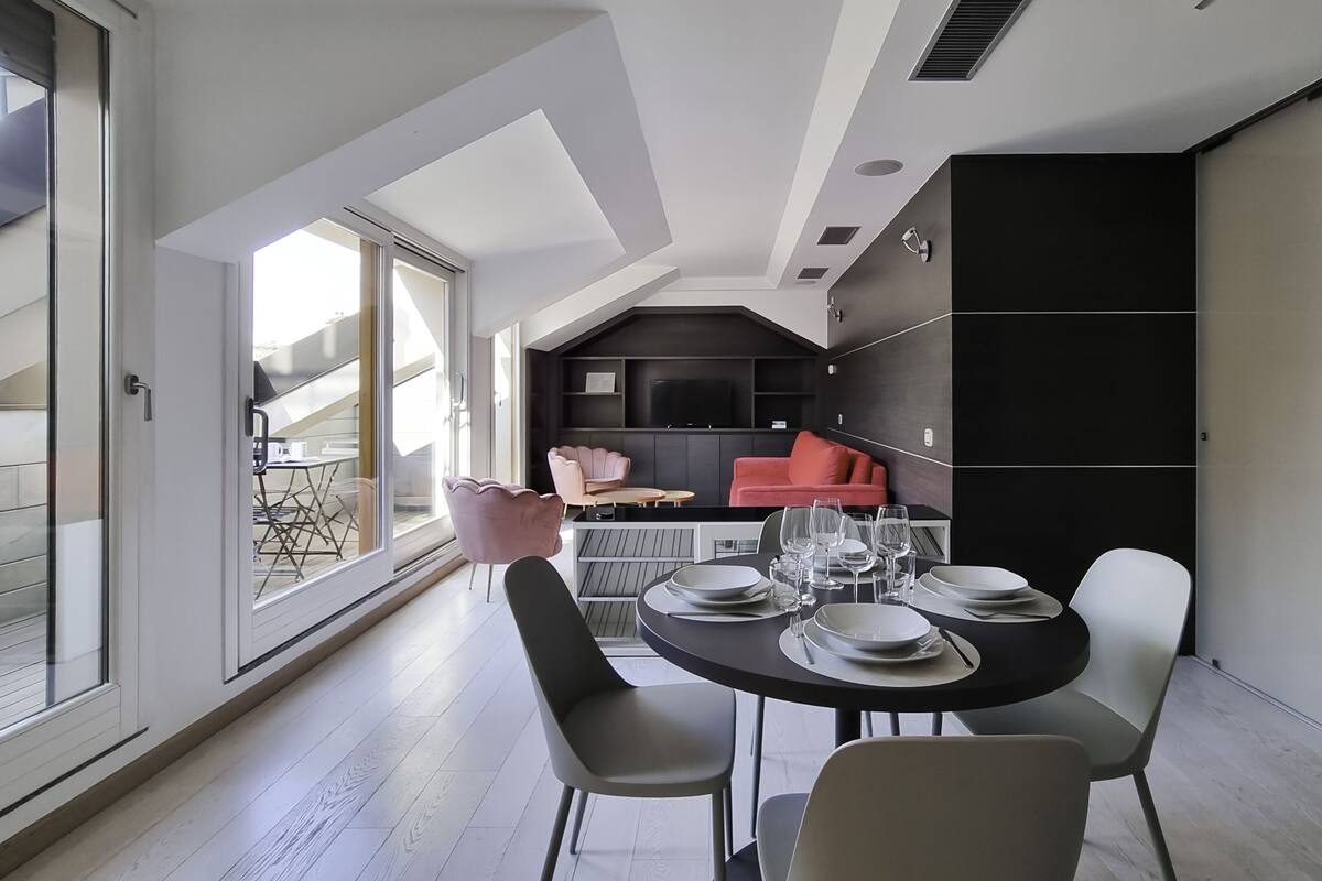 A modern dining area with a round table set for four, a kitchen in the background, and a view of a balcony.