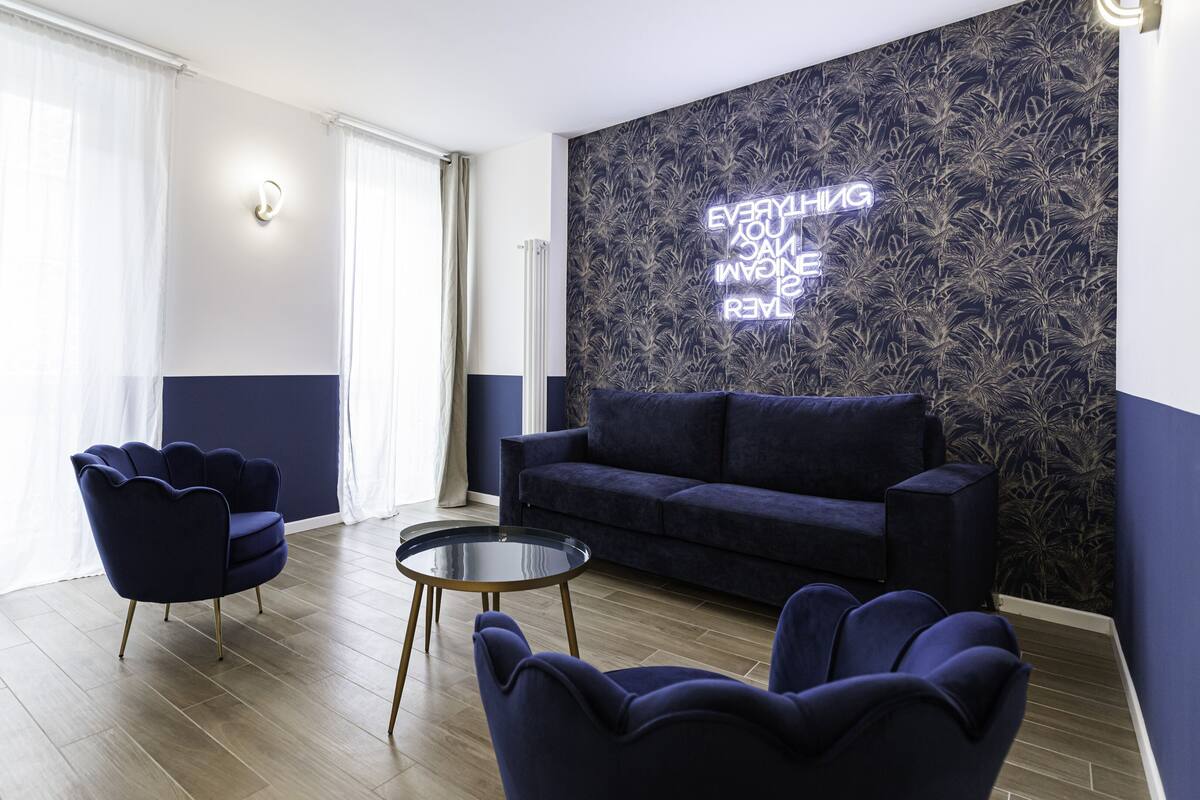 A modern living room with a dark blue sofa, two matching armchairs, a glass coffee table, and a patterned wallpaper with a neon sign.