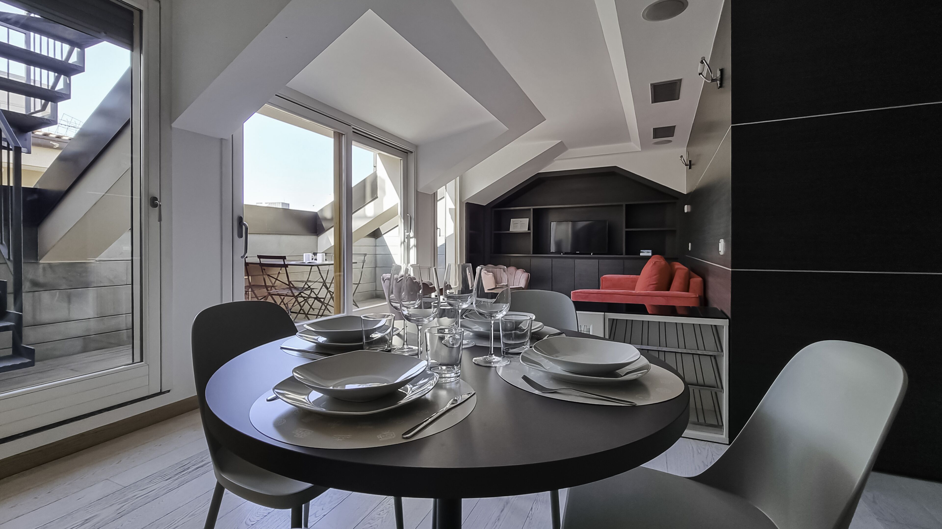 Un coin repas avec une table ronde dressée pour quatre personnes, un espace cuisine avec un canapé rouge et une vue sur le balcon extérieur.