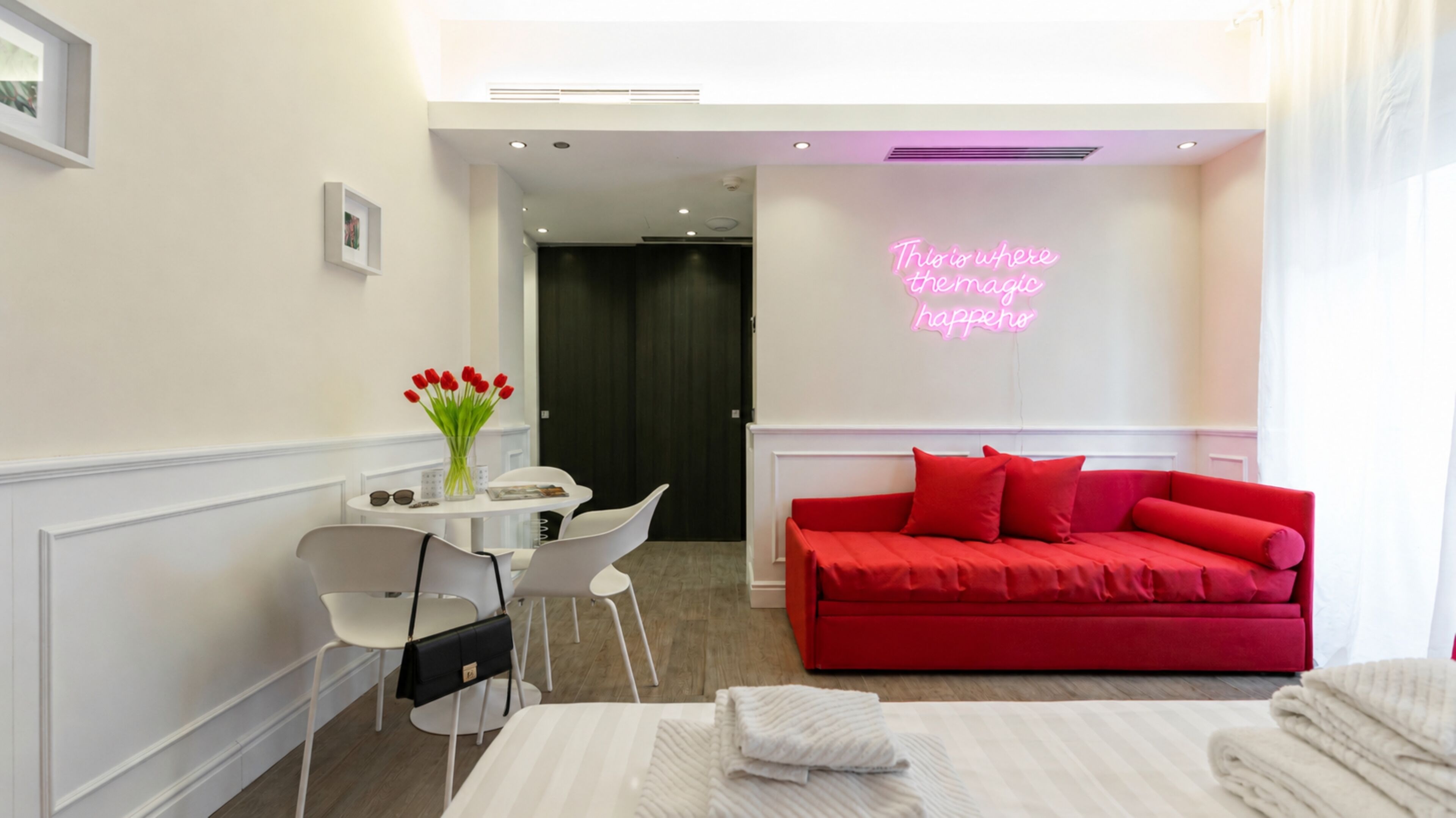 Una habitación de hotel con una cama, un sofá rojo, una mesita con un florero de flores rojas y un letrero de neón en la pared.