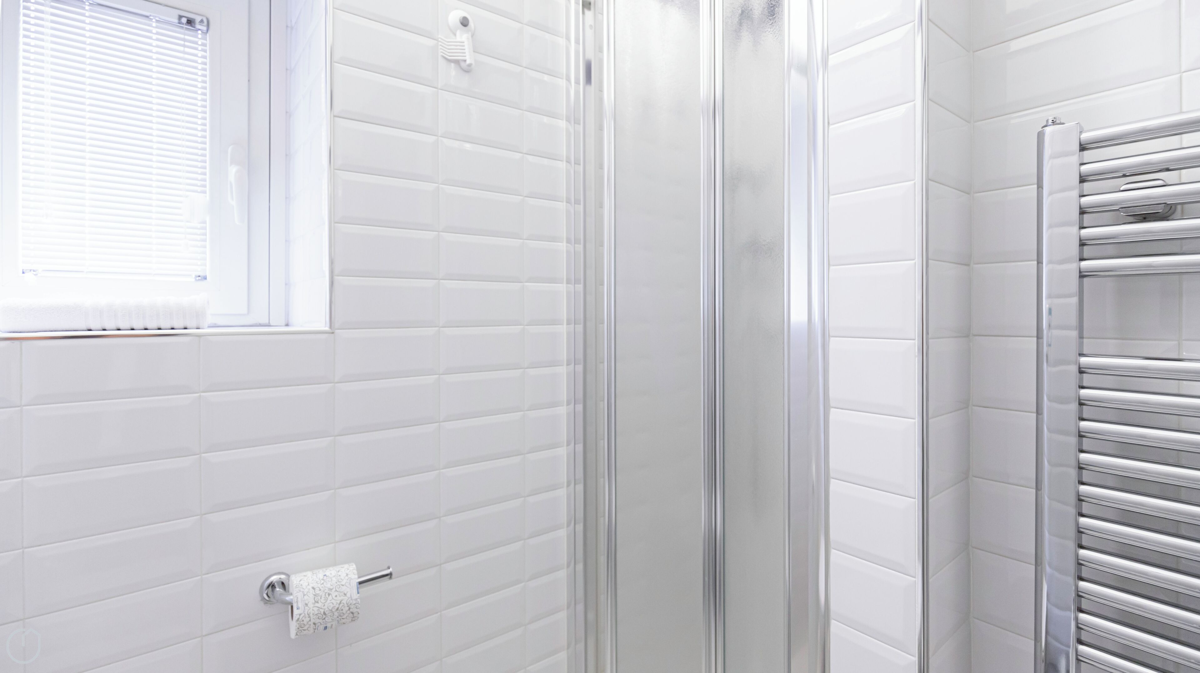 A modern bathroom with white tiled walls, a glass shower enclosure, a towel rack, and a window with blinds.