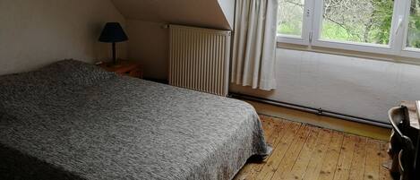 A bedroom with a sloped ceiling, a bed with a textured blanket, a bedside lamp, a radiator, and a window with curtains.