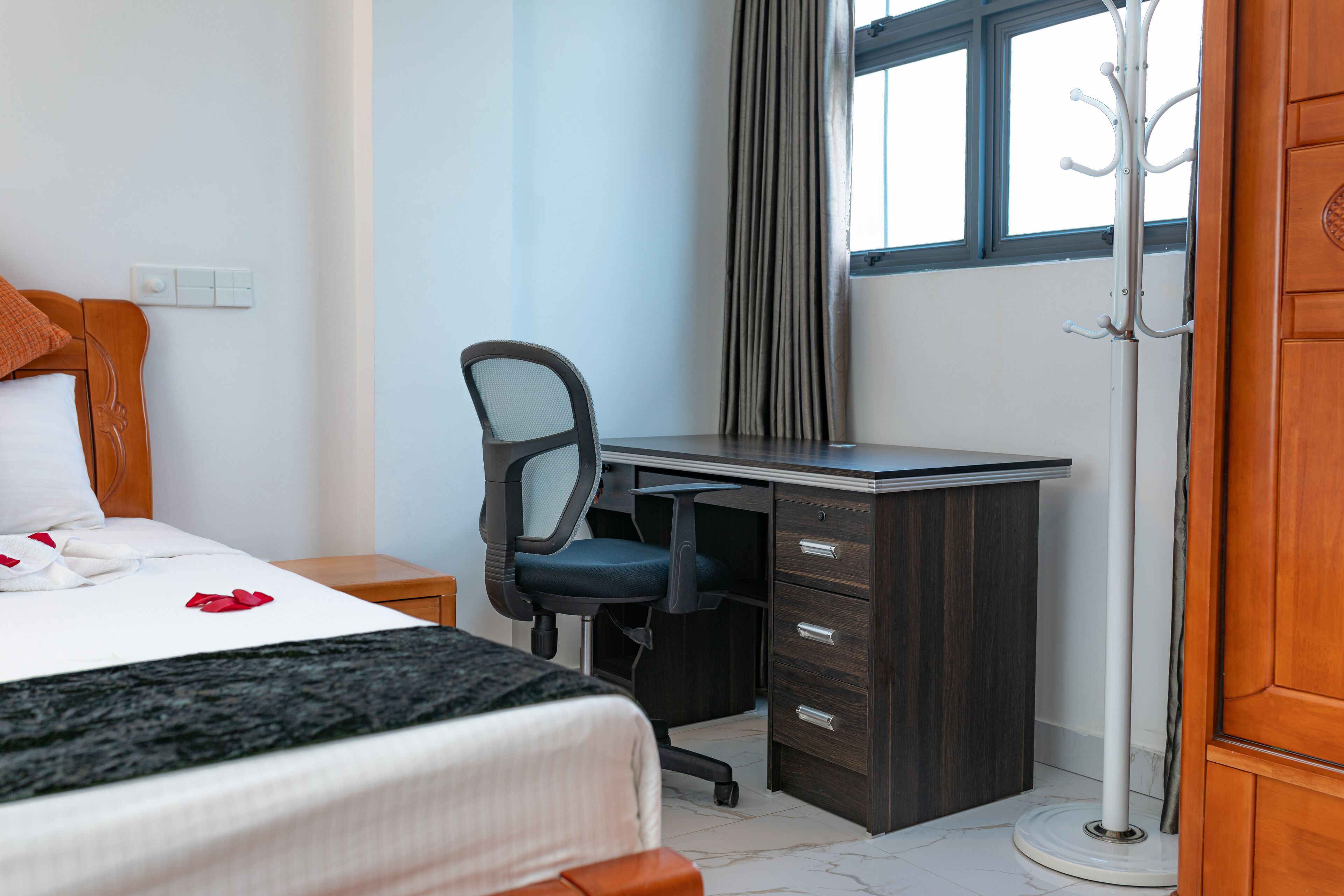 A hotel room with a bed, a desk, a chair, a coat rack, and a window.