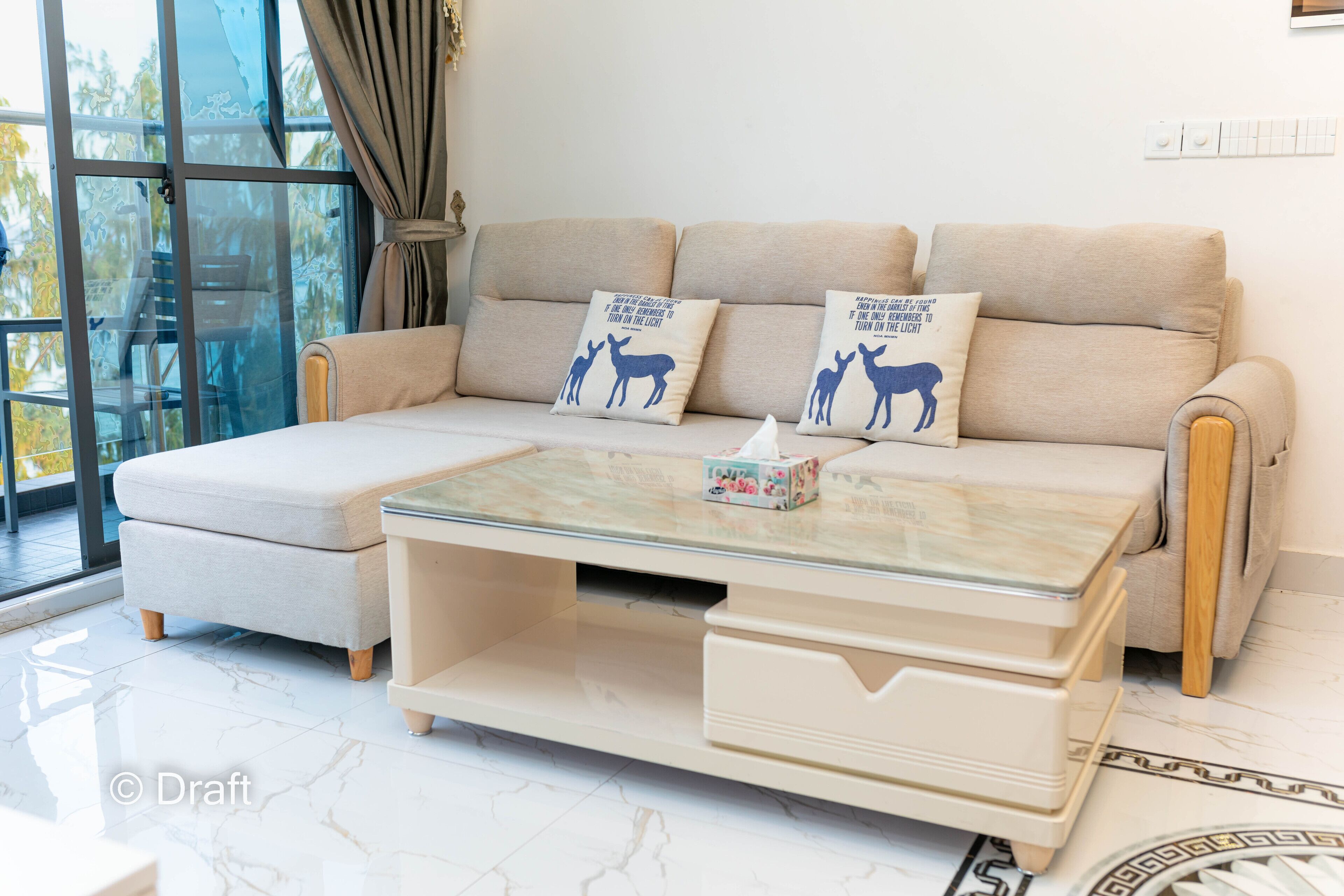 A modern living room with a beige sofa, two decorative pillows, a glass coffee table, and a view through large windows.