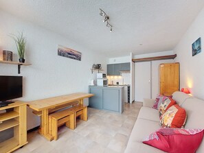 A compact living space with a wooden dining table, a sofa with colorful cushions, a television, a kitchen area with a microwave and stove, and a small plant on a shelf. - Studio for 4 pers with south balcony near shops and slopes (Montvalezan-La Rosière)