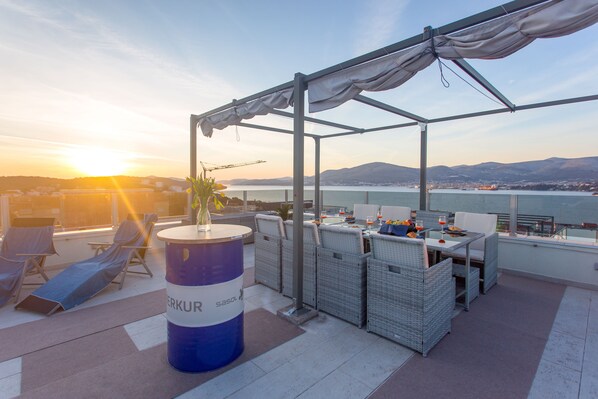 A rooftop terrace with a table and chairs, a large barrel, and a view of the sea and mountains.