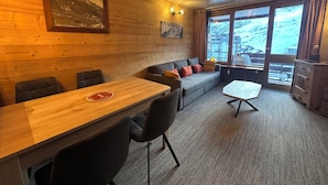 Une pièce chaleureuse avec un plafond en bois, un canapé, une table et des chaises. Des tableaux ornent les murs et de grandes fenêtres offrent une vue sur l’extérieur.