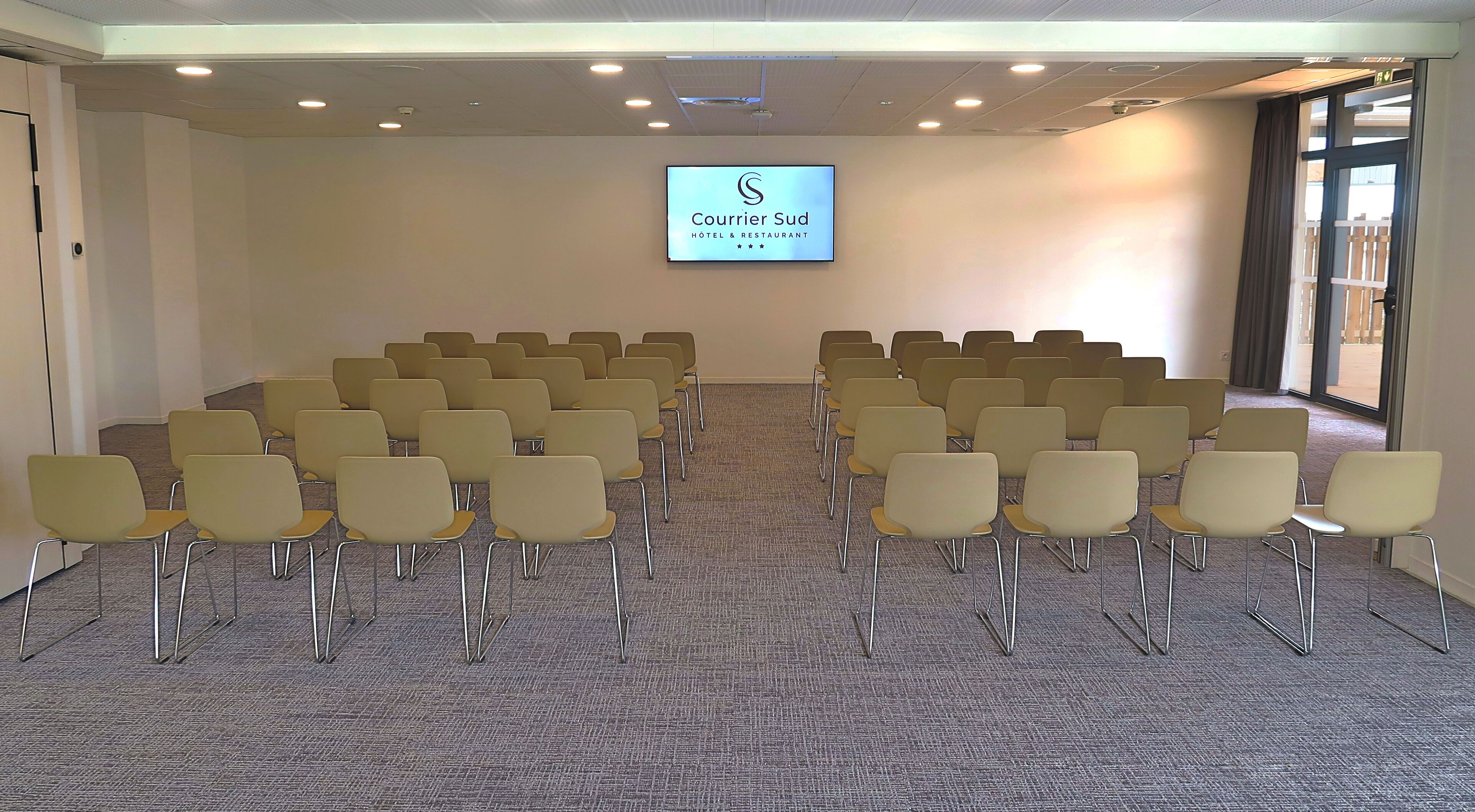 Un salón de conferencias con filas de sillas, una pantalla de proyección mostrando 'Courrier Sud Hotel & Restaurant' y vistas al exterior a través de un amplio ventanal.