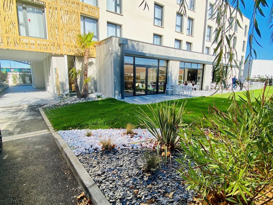 Ein modernes Gebäude mit Glastür, ein angelegter Garten mit Steinen und Pflanzen sowie ein klarer, blauer Himmel.