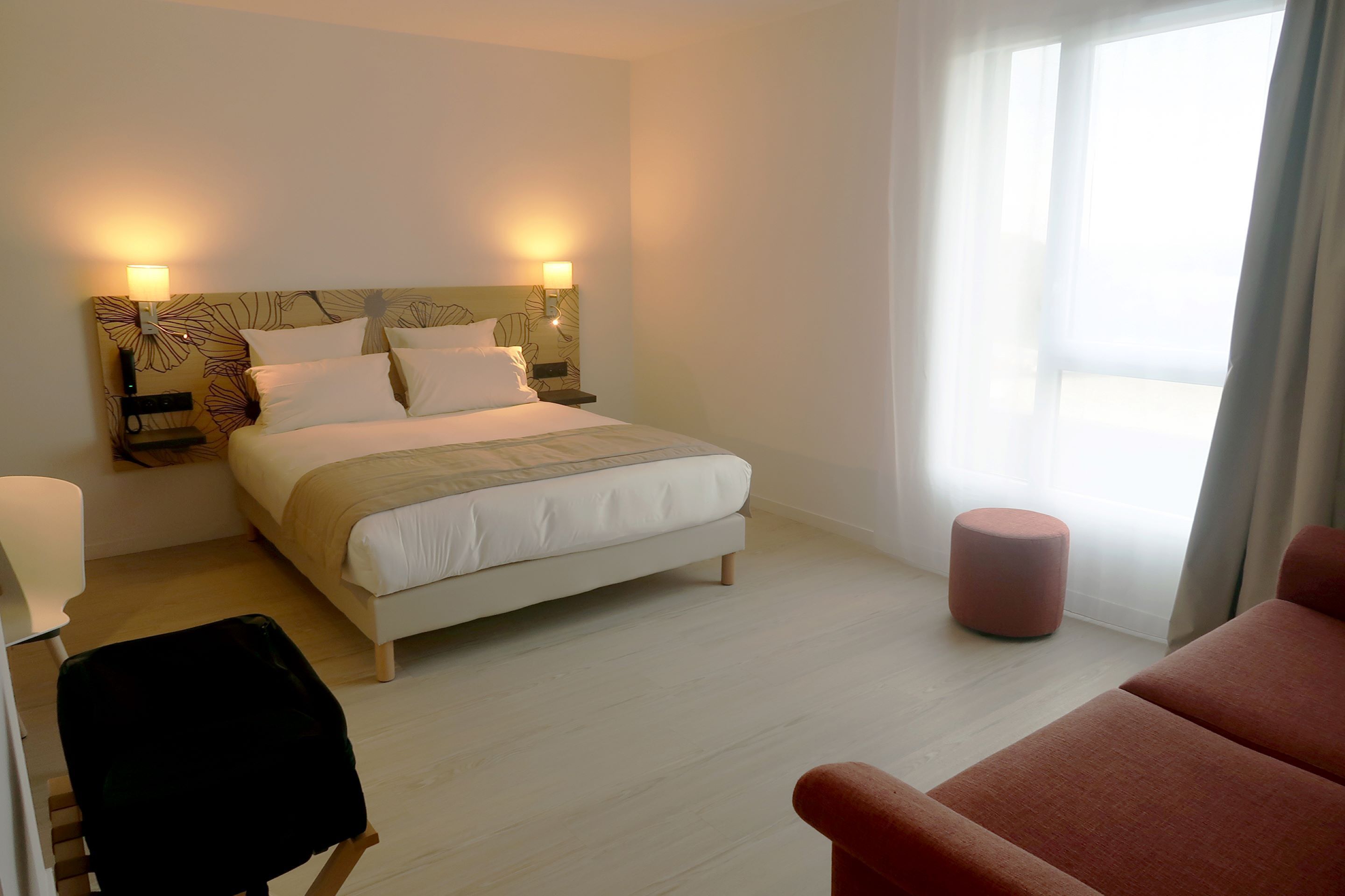 A hotel room with a bed, bedside lamps, a chair, a small ottoman, and a view of the outdoors.