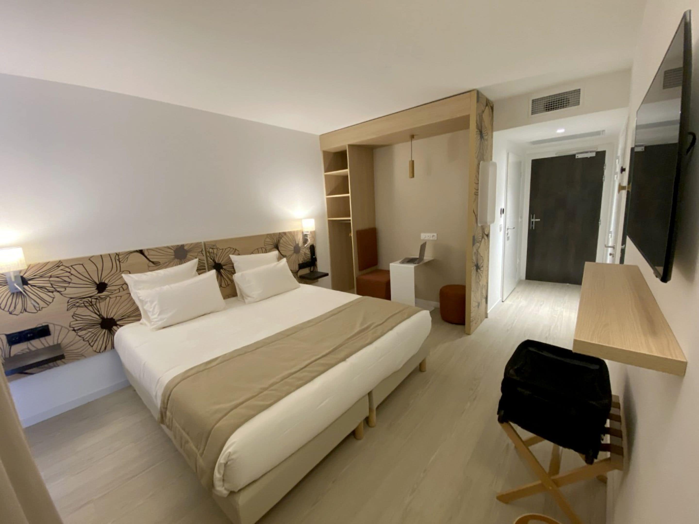 A hotel room with a large bed, a desk, a chair, a TV, and a wardrobe.
