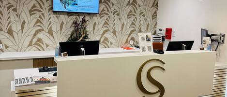 A modern reception desk with a large 'S' logo, a mounted TV screen, and a patterned wallpaper background.