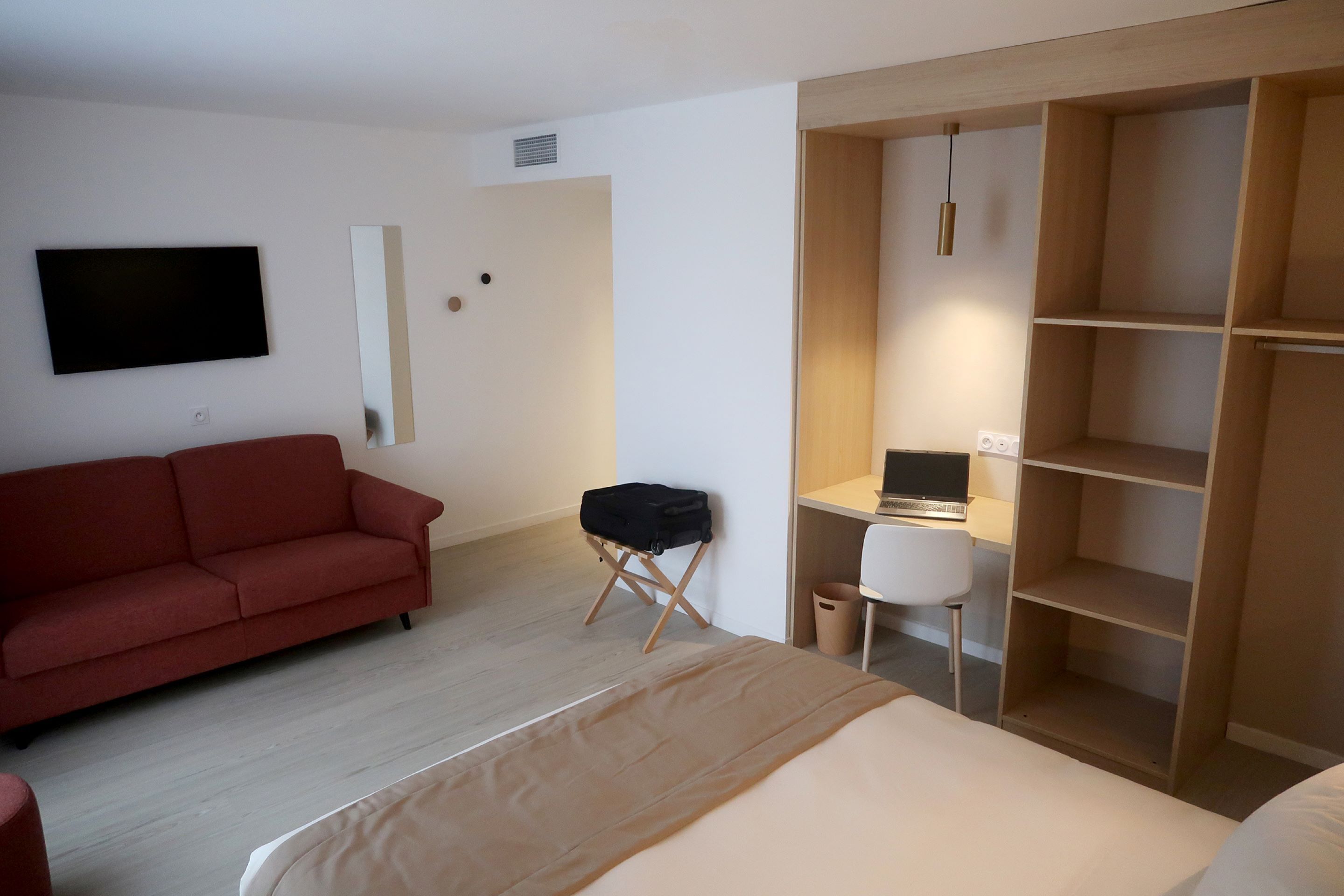 A modern hotel room with a bed, a red sofa, a desk with a computer, and a wardrobe.