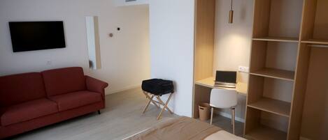 A modern hotel room with a bed, a red sofa, a desk with a computer, and a wardrobe.