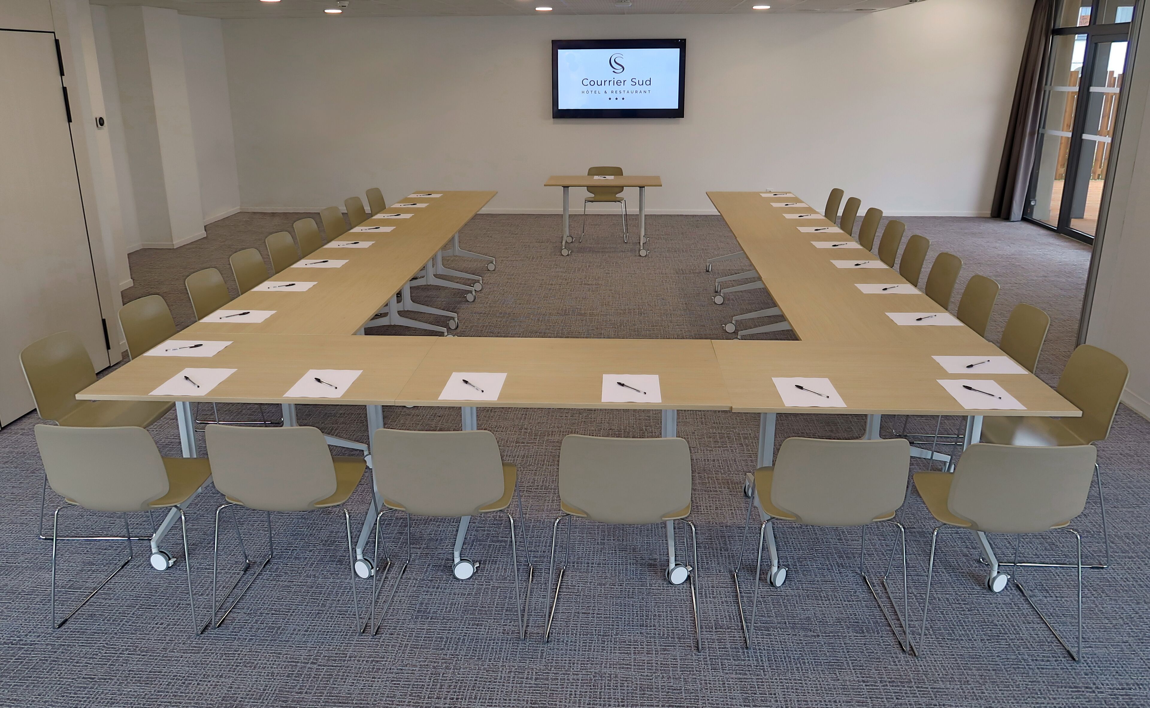 Un salón de conferencias con una larga mesa, sillas, una pantalla de proyector y piso alfombrado.