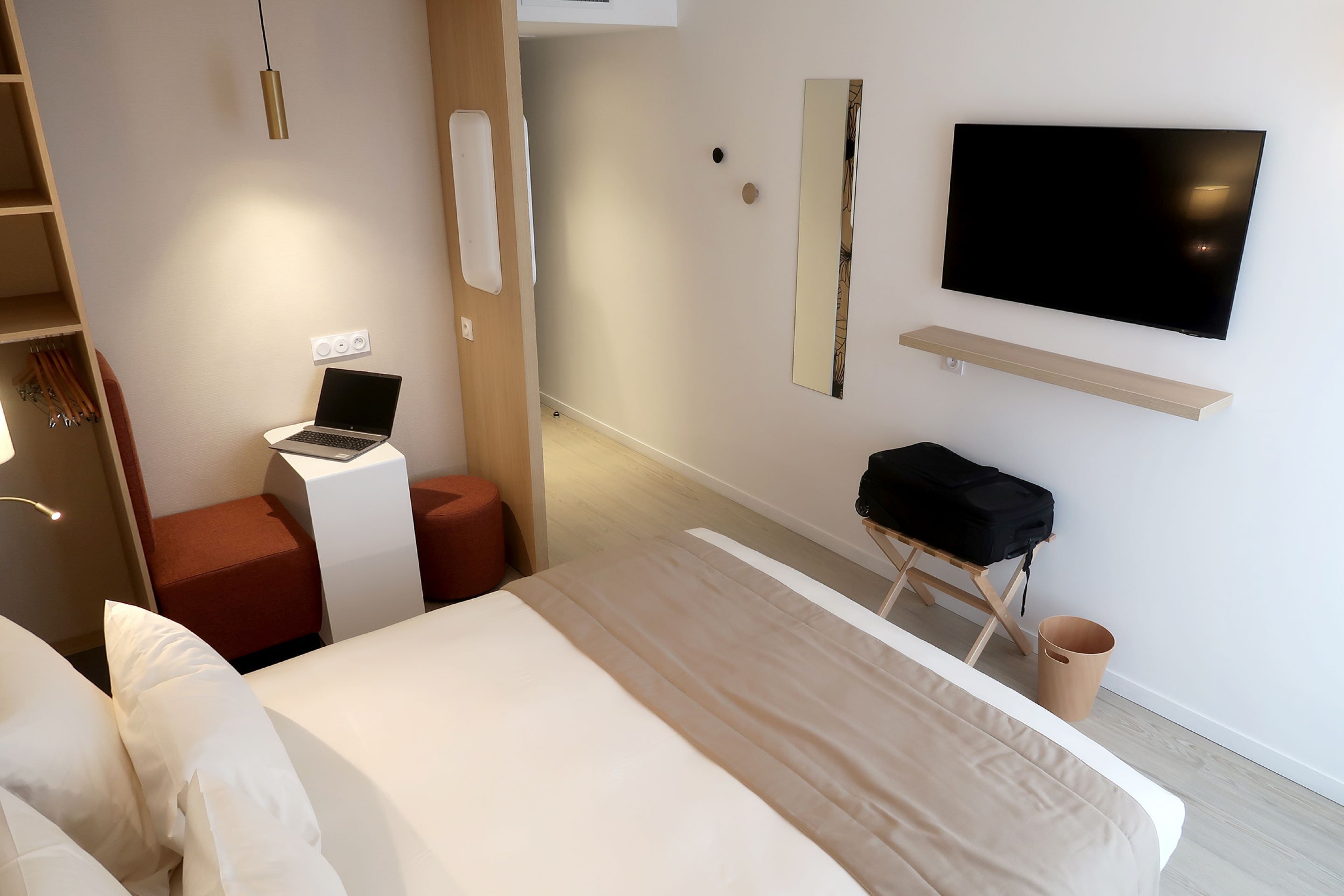 A modern hotel room with a bed, a desk with a laptop, a TV mounted on the wall, and a small seating area.