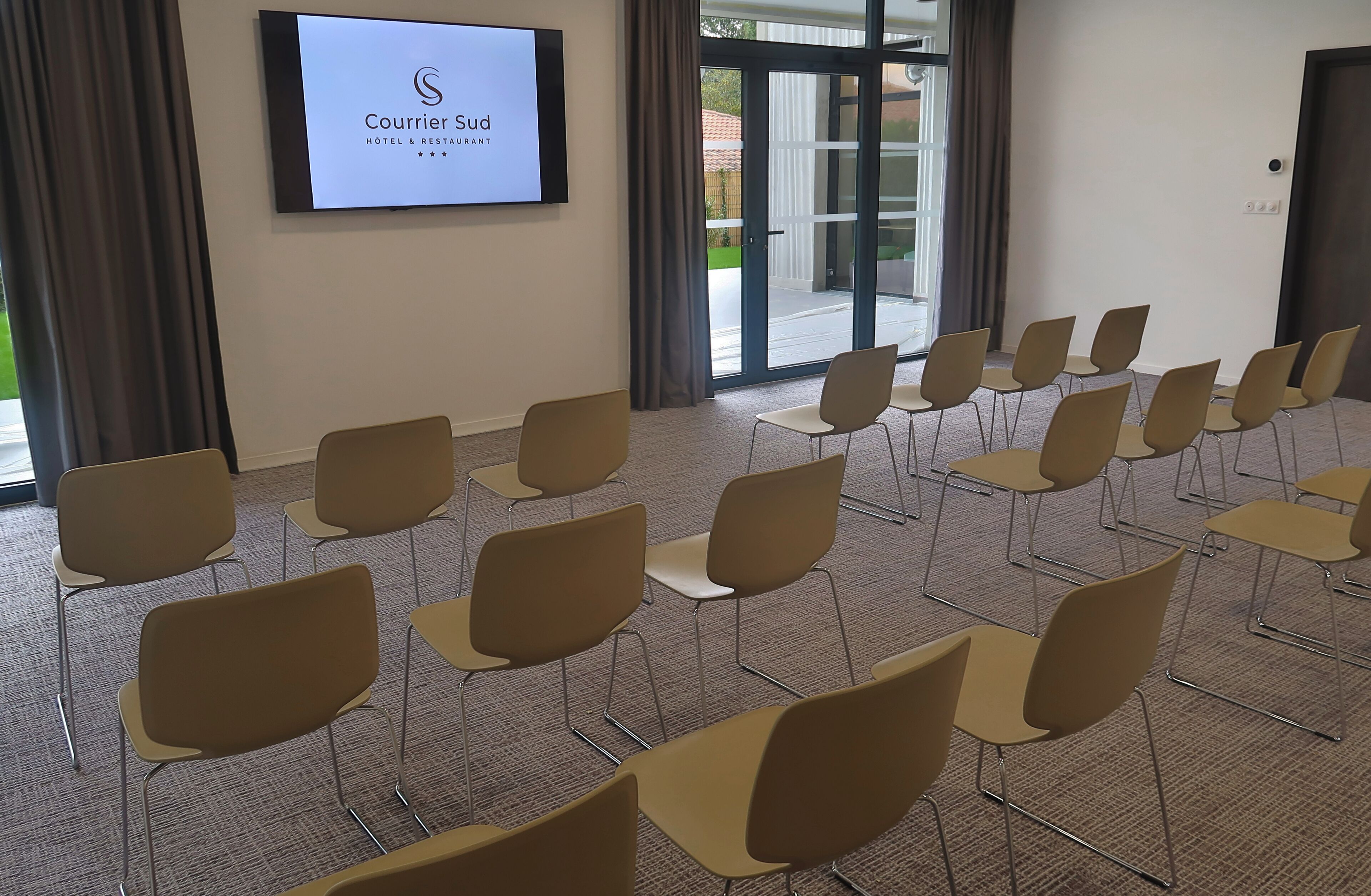 Un salón de conferencias con filas de sillas, una pantalla grande mostrando el logotipo del Hotel & Restaurante Courrier Sud, y vista del área exterior a través de puertas de vidrio.
