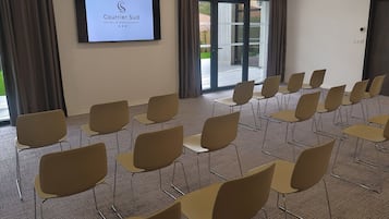 A conference room with rows of chairs, a large screen displaying the Courrier Sud Hotel & Restaurant logo, and a view of the outdoor area through glass doors.