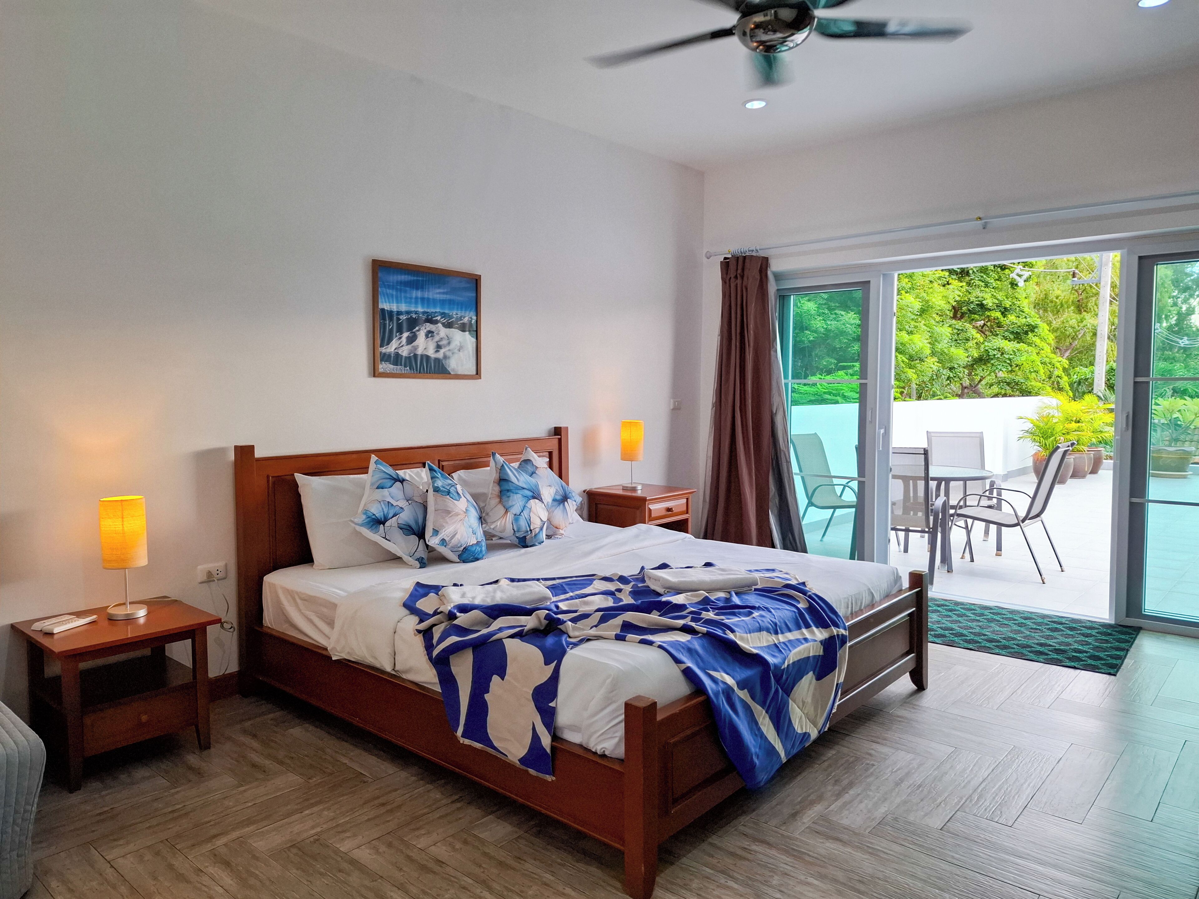 A hotel room with a bed, bedside tables, a ceiling fan, a window with curtains, and a view of greenery outside.