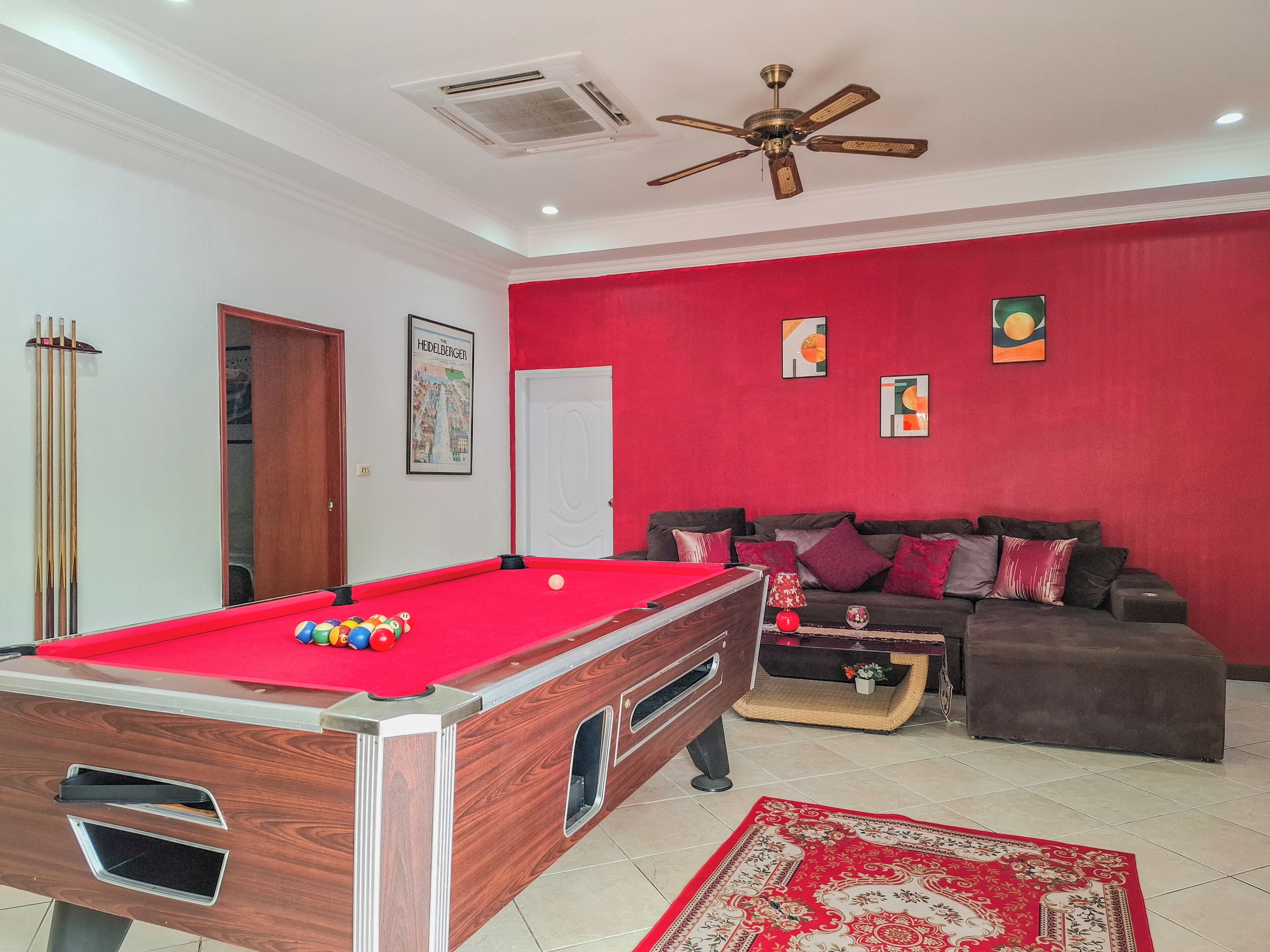 A room with a pool table, a sofa, a ceiling fan, and a red wall.