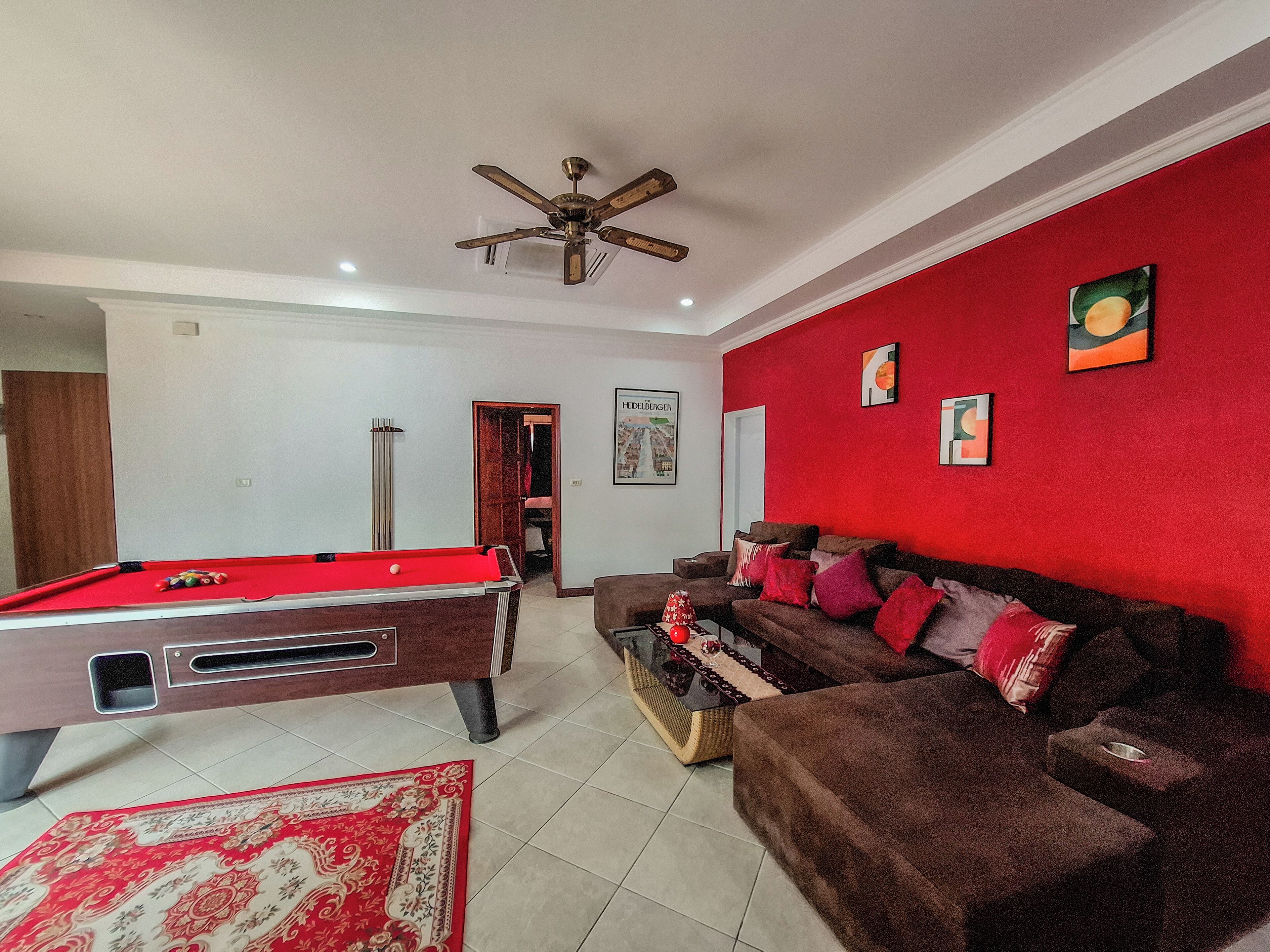 Un salon avec une table de billard, un canapé marron et un mur d’accent rouge.