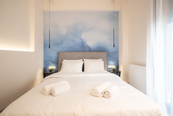 Un lit bien fait, avec du linge de lit blanc et deux serviettes enroulées, une tête de lit rembourrée de couleur grise et un mur orné d’un tableau abstrait bleu.