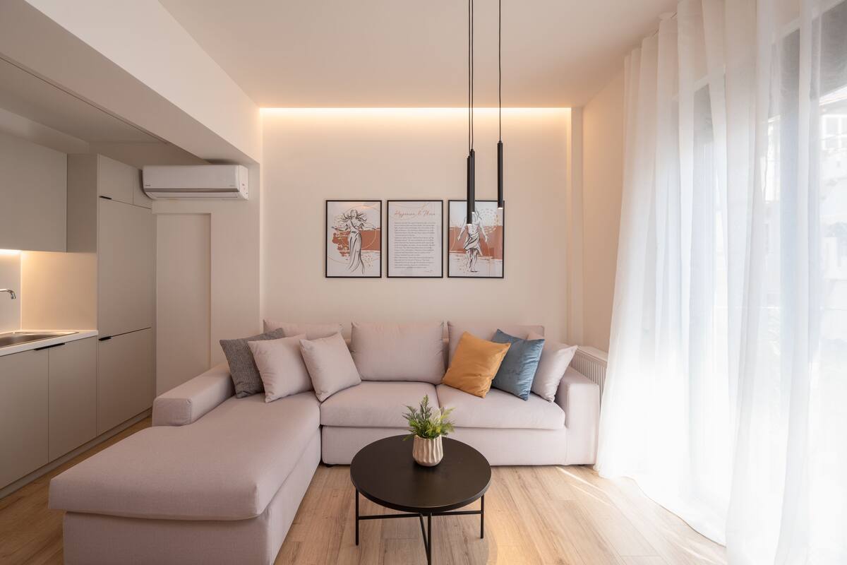 A modern living room with a sofa, a coffee table, and a kitchen area. There are three framed artworks on the wall.